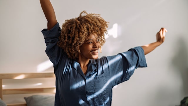 Beautiful woman sitting in her bed in silk pajamas. She is stretching with her arms up in the air and smiling. Sun rays are coming in through the blinds.