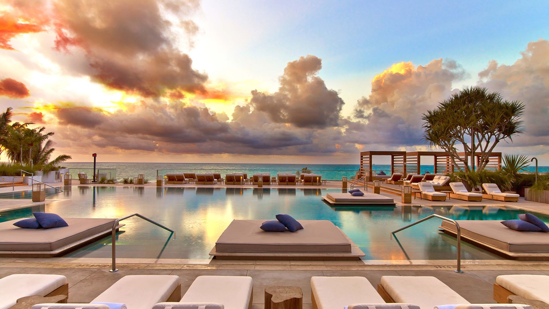 A luxurious oceanfront infinity pool at sunset, surrounded by cushioned sun loungers and floating daybeds with navy pillows. The scene is bathed in golden and pastel hues from the dramatic sky, with palm trees and lush greenery framing the serene setting.