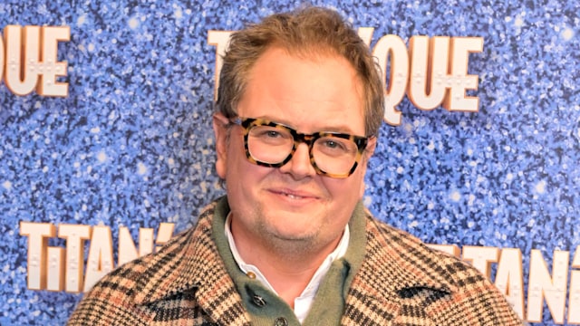 Alan Carr wearing checkered coat standing in front of 'Titanique' step and repeat at the West End opening