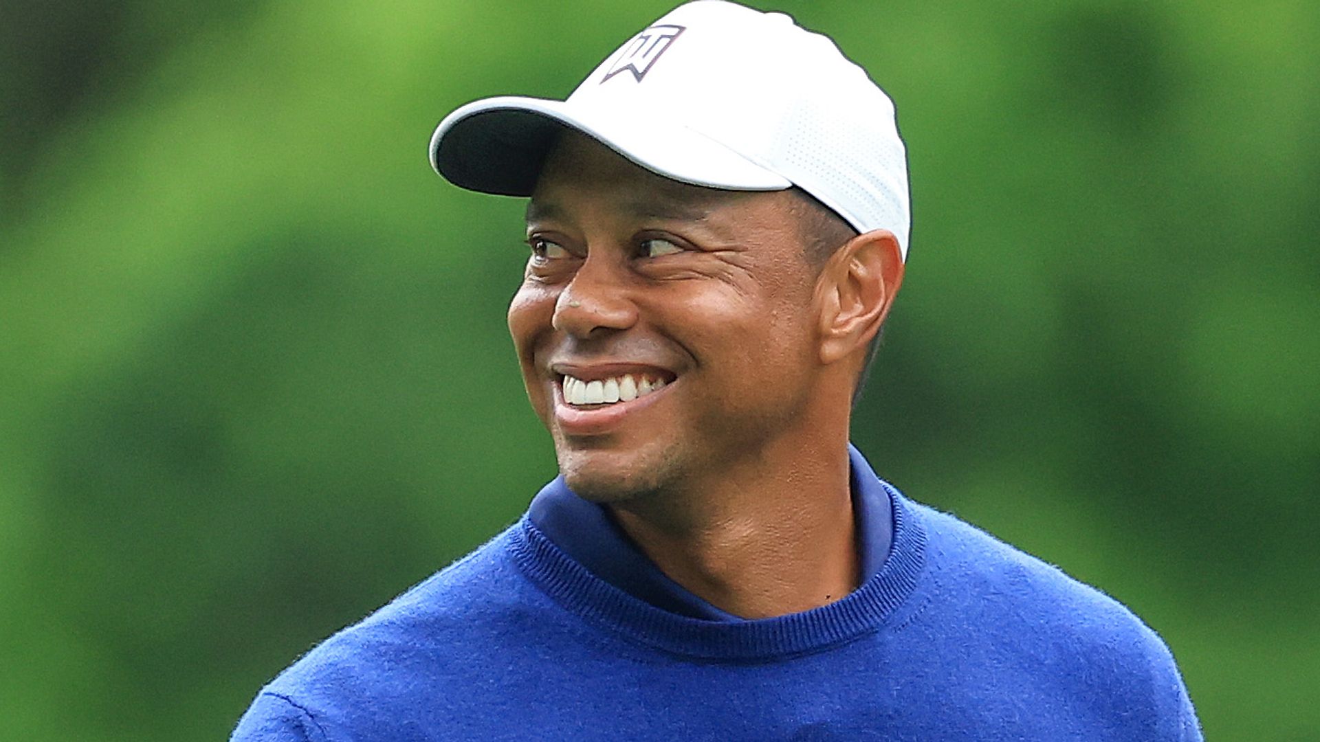 Tiger Woods smiling in a blue jumper with a golf club in hand