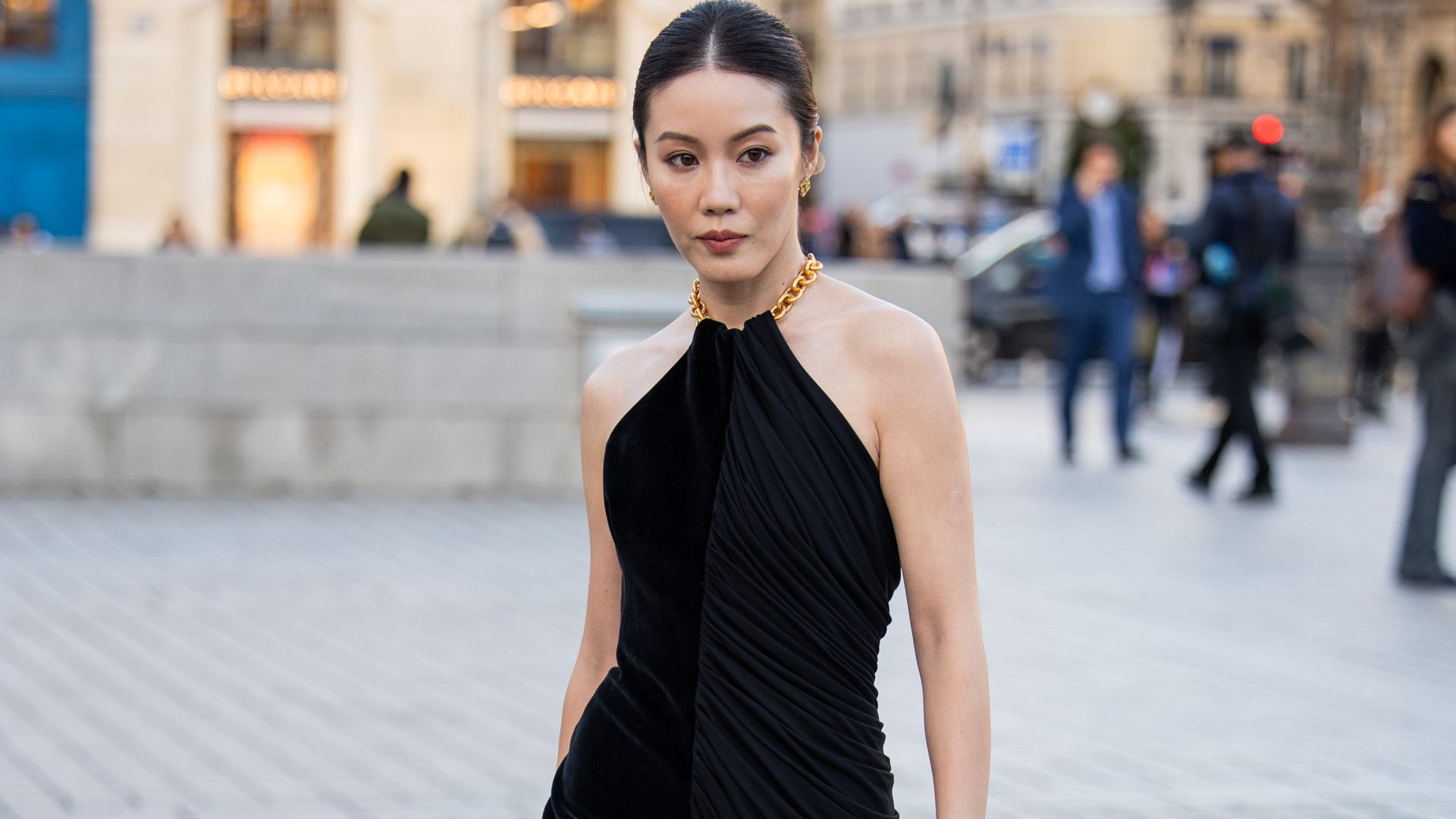 Jenny Tsang wears black dress with slit, golden bag outside Schiaparelli during Womenswear Spring/Summer 2025 as part of Paris Fashion Week
