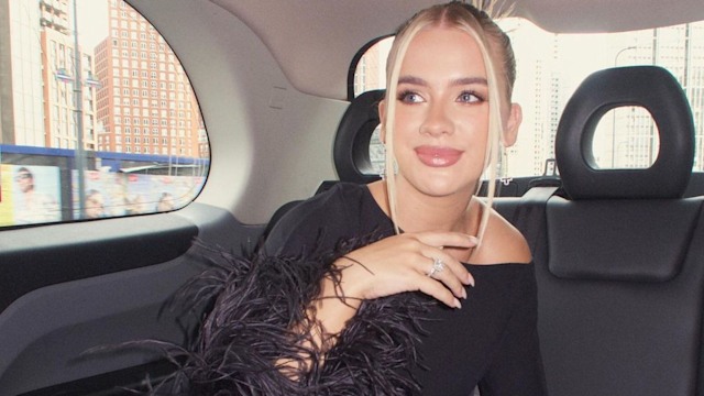 Grace Beverley sitting in the back of a car. She is wearing an elegant black outfit with feather details and her hair styled neatly in an updo with loose strands framing her face. She looks glamorous, exuding a confident and poised expression while looking off to the side.