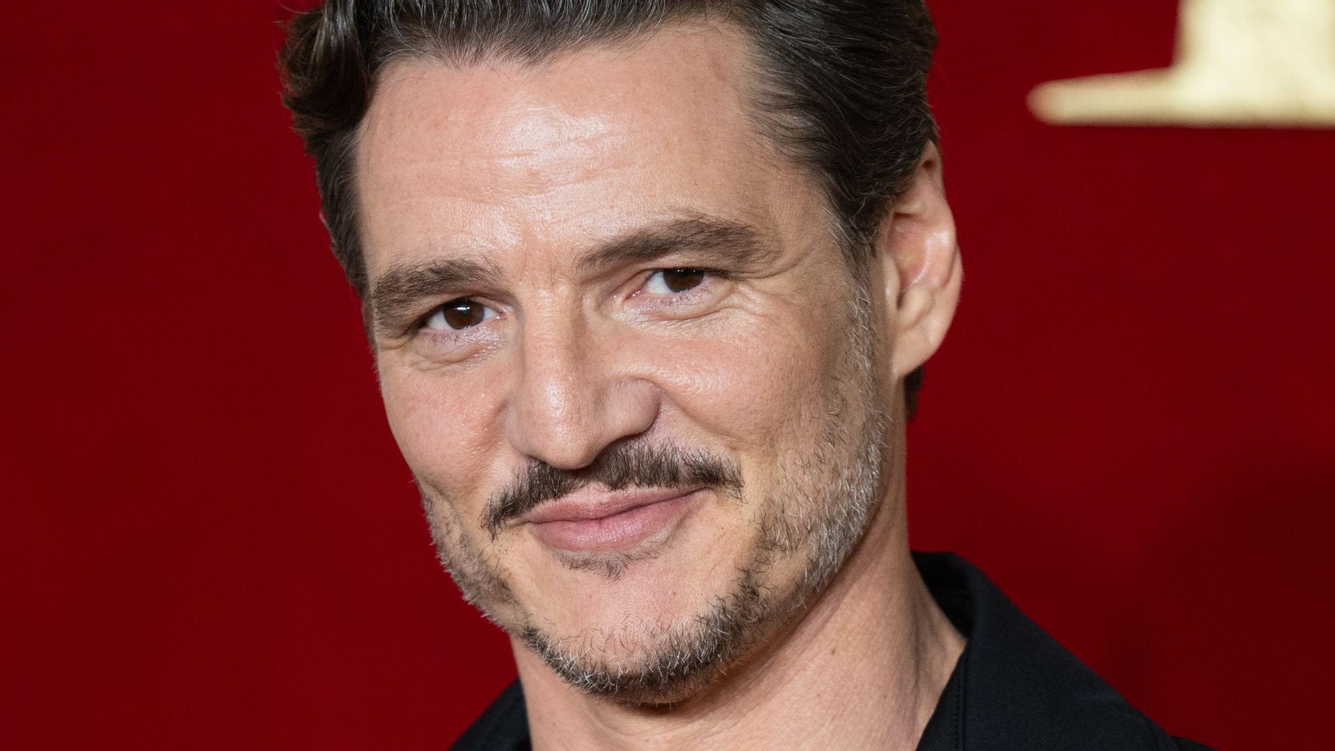 Pedro Pascal in a low plunging black shirt with red detailing, sporting a moustache and coiffed hair. 