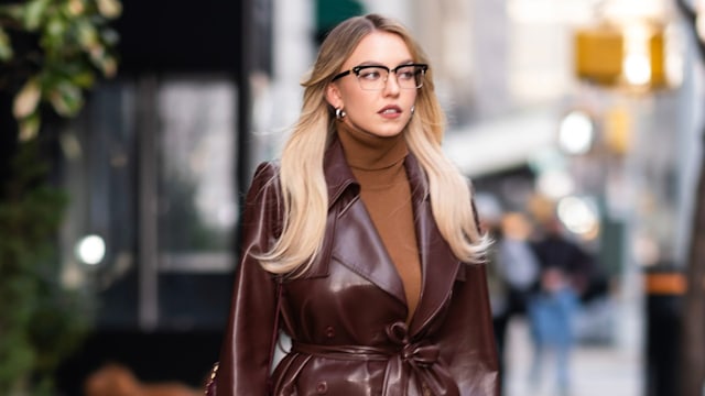 Sydney Sweeney wears brown leather trench coat with brown turtle neck and nerdy glasses while walking down sidewalk in new york