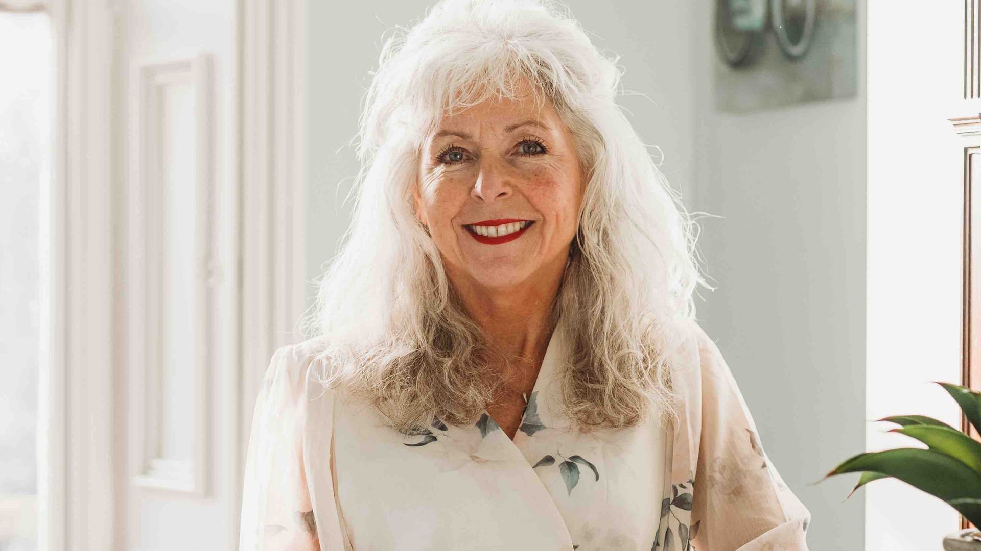 Woman with grey hair in a white blouse posing next to a fireplace