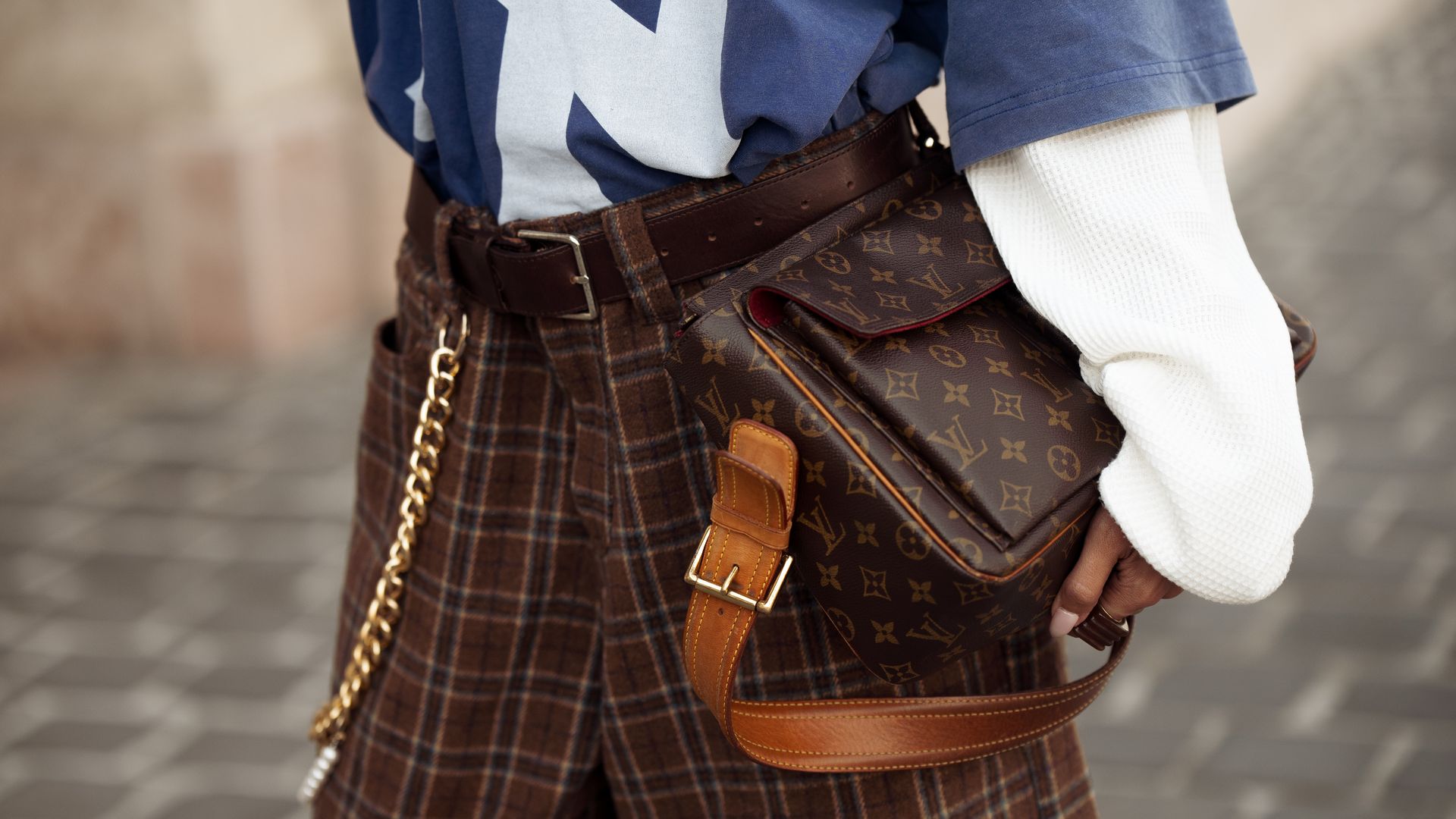 A guest wears brown checkered shorts, white long sleeve, blue NY top and brown Louis Vuitton bag during the Budapest Central European Fashion Week