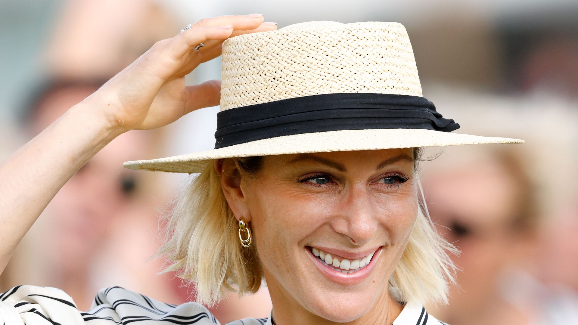Zara Tindall holds boater hat on head smiling in striped dress