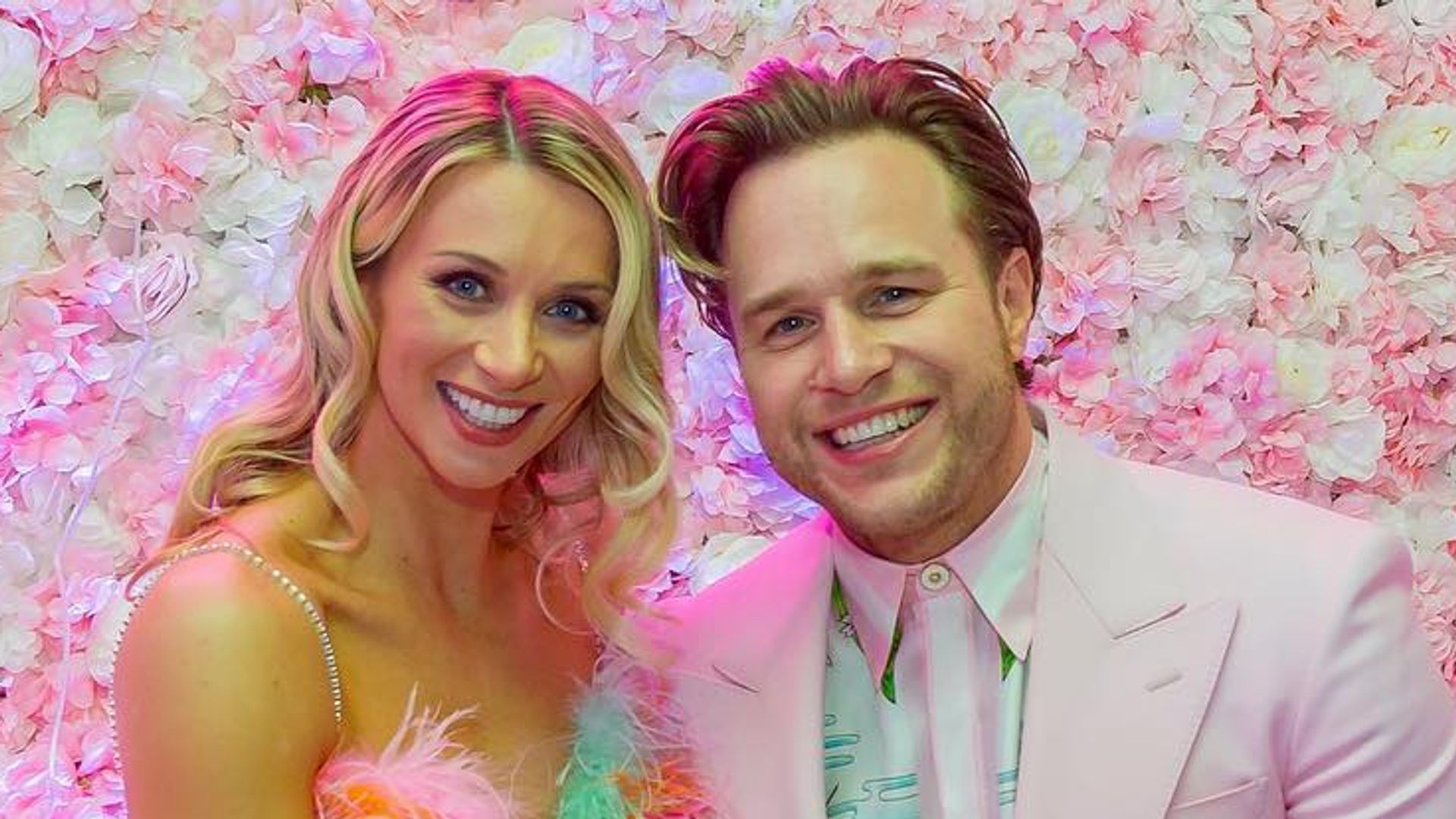 Olly Murs posing with feather dress clad fiancee Amelia Tank in front of flower wall