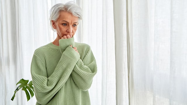Middle-aged woman with gray hair nervously biting her nails while standing at home window