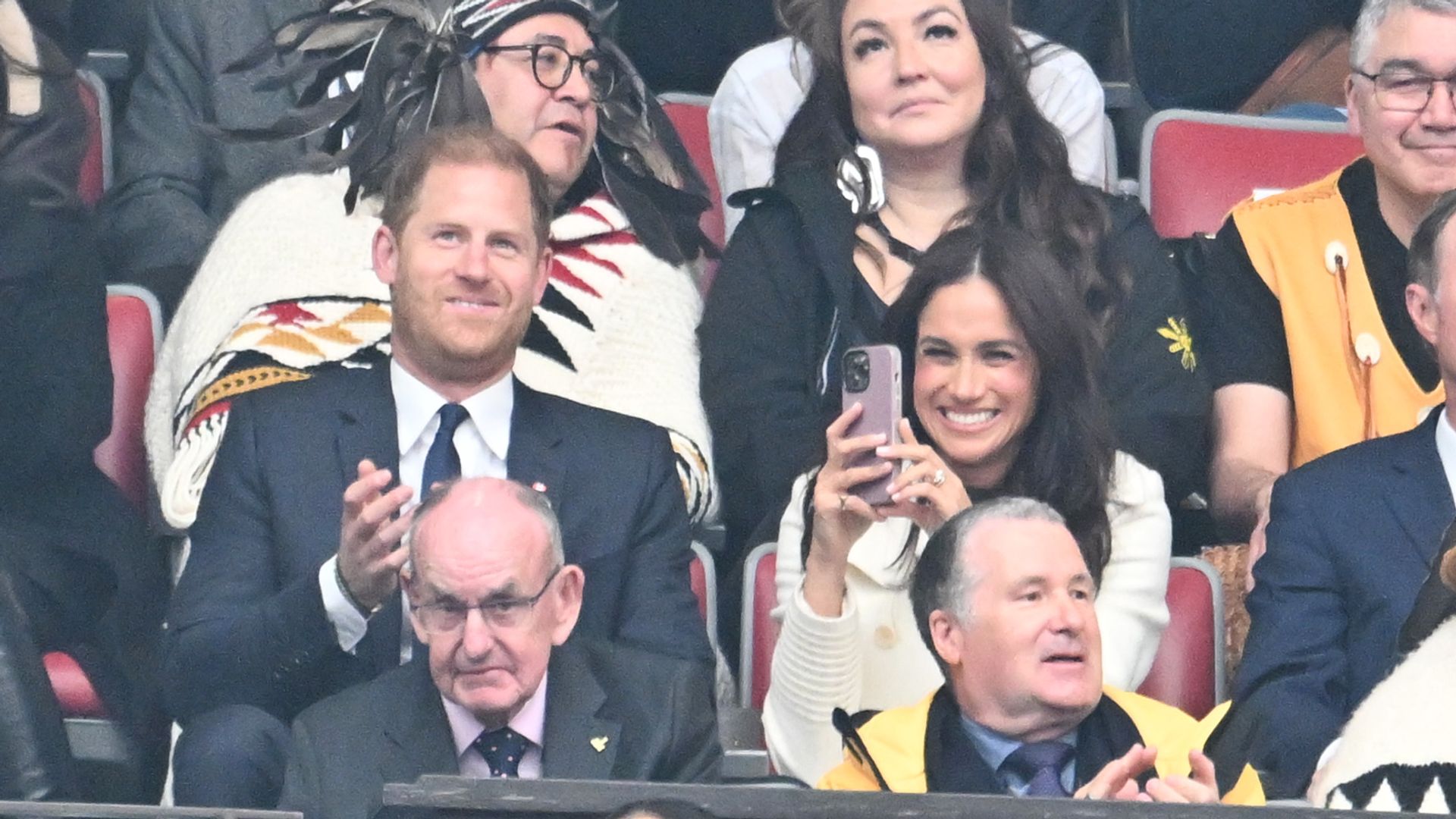 Prince Harry applauding and Meghan Markle recording on a phone