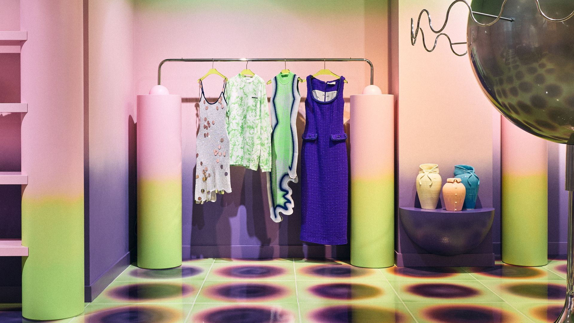 A vibrant KOIBIRD clothing display with pastel-gradient walls, sculptural racks, and a futuristic chrome orb centerpiece surrounded by bold printed tiles.