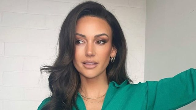 Michelle Keegan in green dress with brunette curly hair posing against white wall