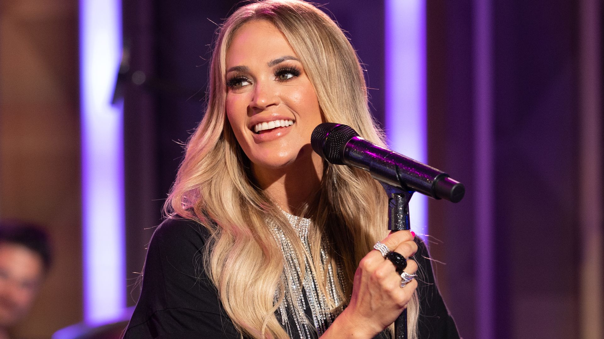 Carrie Underwood smiling while looking to the left, she is standing with a mic stand in front of her