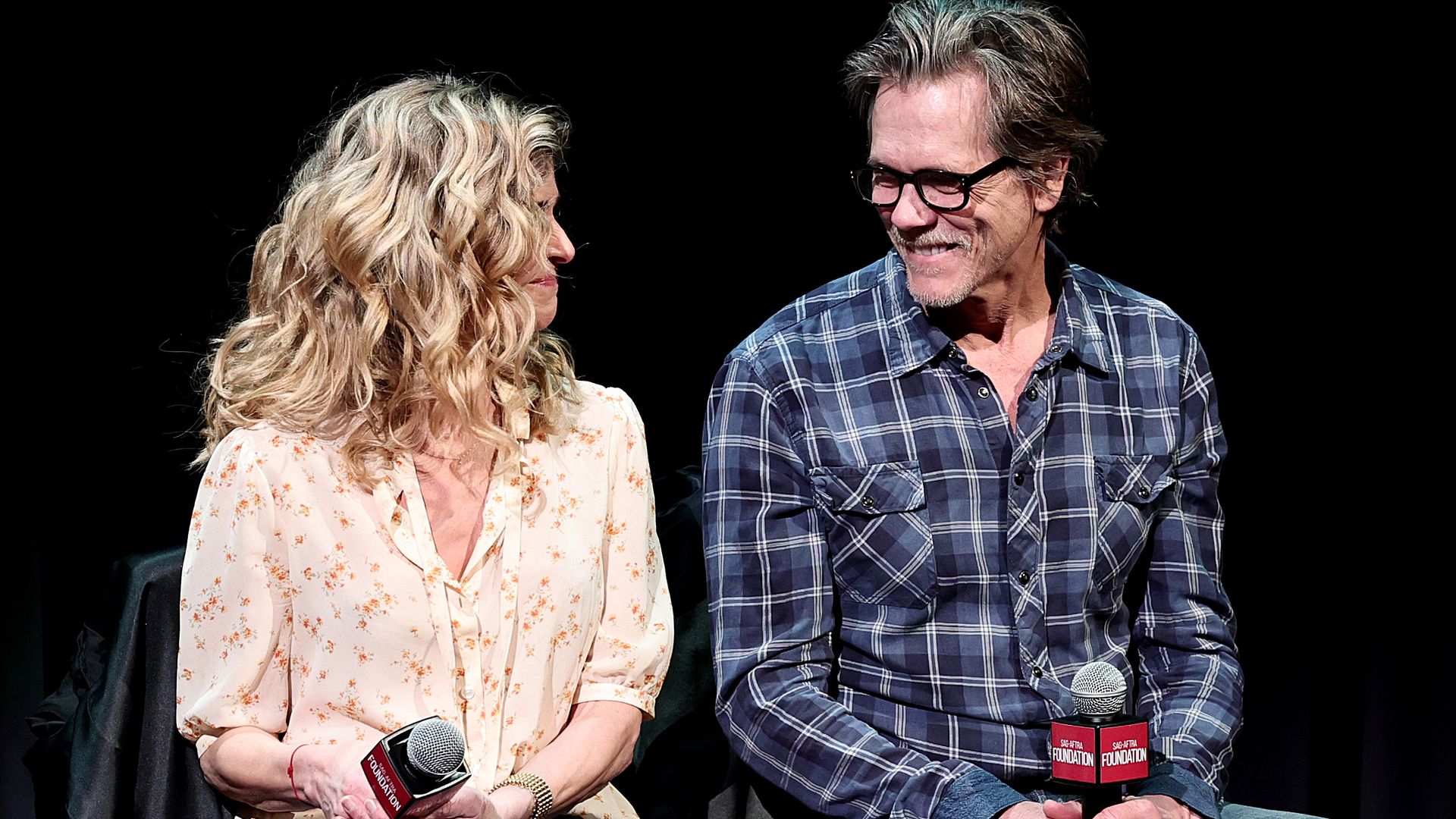 Kevin Bacon and Kyra Sedgwick smiling at each other while sat on stage being interviewed