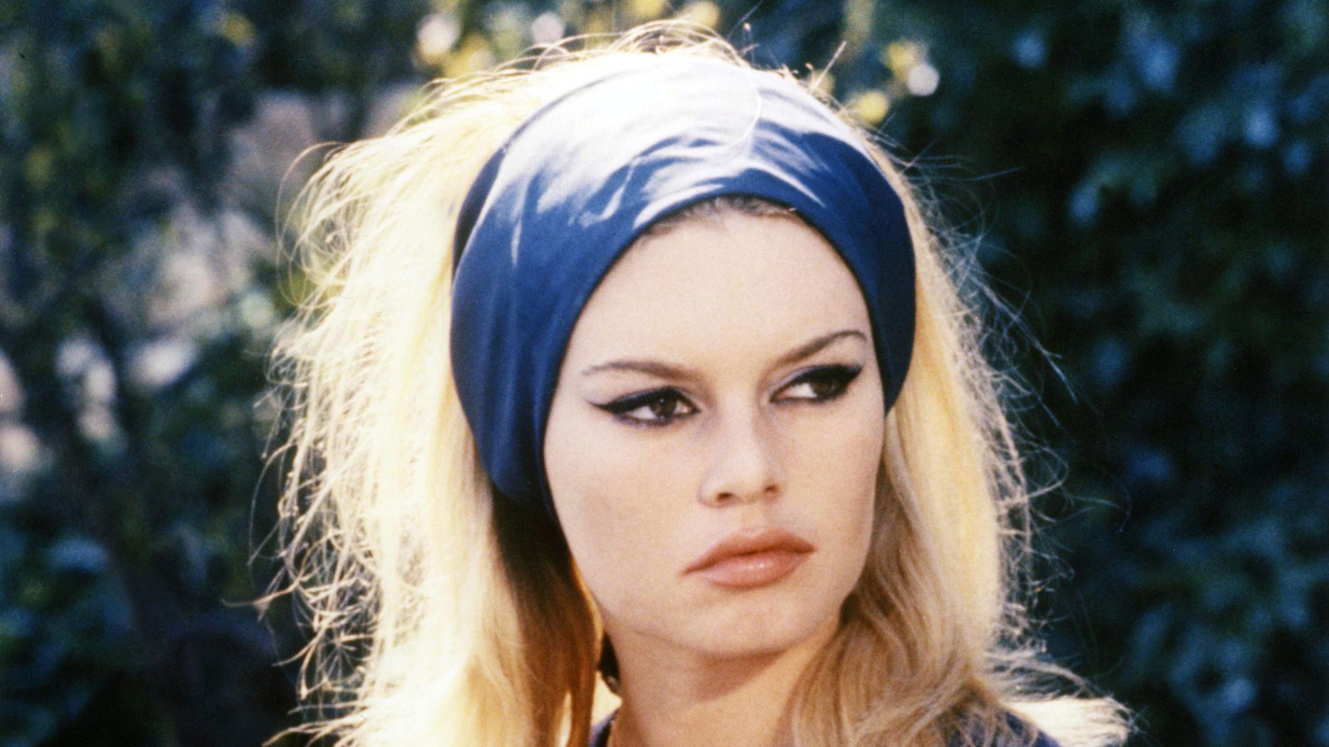 Brigitte Bardot wears a navy headband, blue cardigan and blue and white striped top. Her hair is blonde and bouffant and she has her signature thick black eyeliner on her eyes.