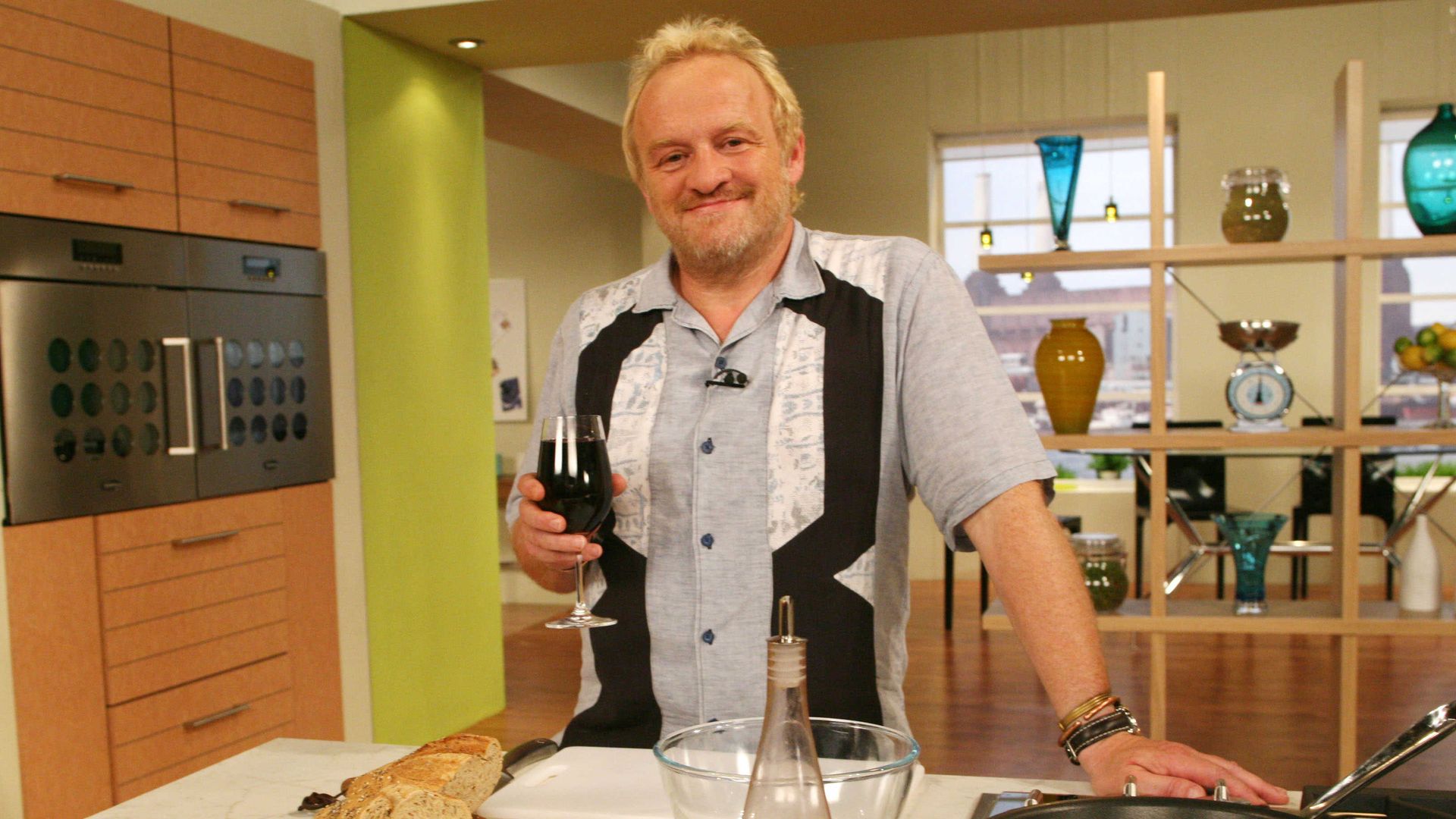 Antony Worrall Thompson on the Saturday Kitchen behind a kitchen counter holding a glass of red wine