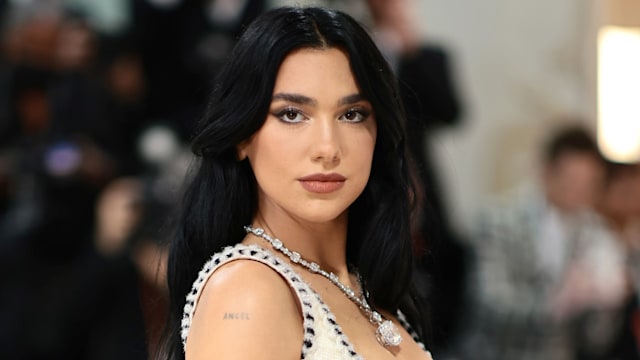 Dua Lipa stands elegantly on the Met Gala red carpet wearing an ivory Chanel gown with black trim, accessorized with a sparkling diamond necklace, her hair styled in loose waves, and “ANGEL” tattoo visible on her upper arm.