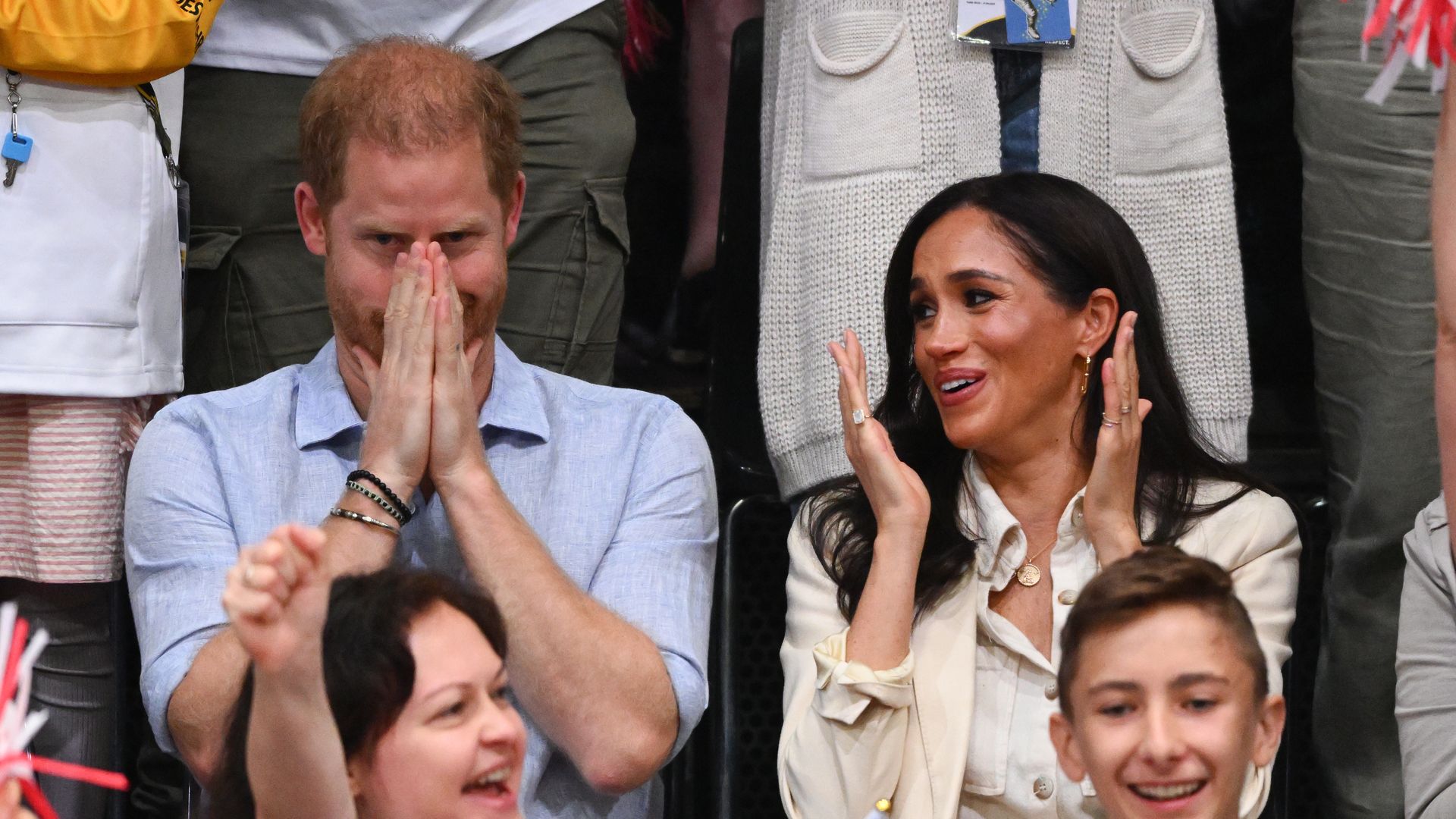 Prince Harry covering his mouth as Meghan Markle applauds next to him