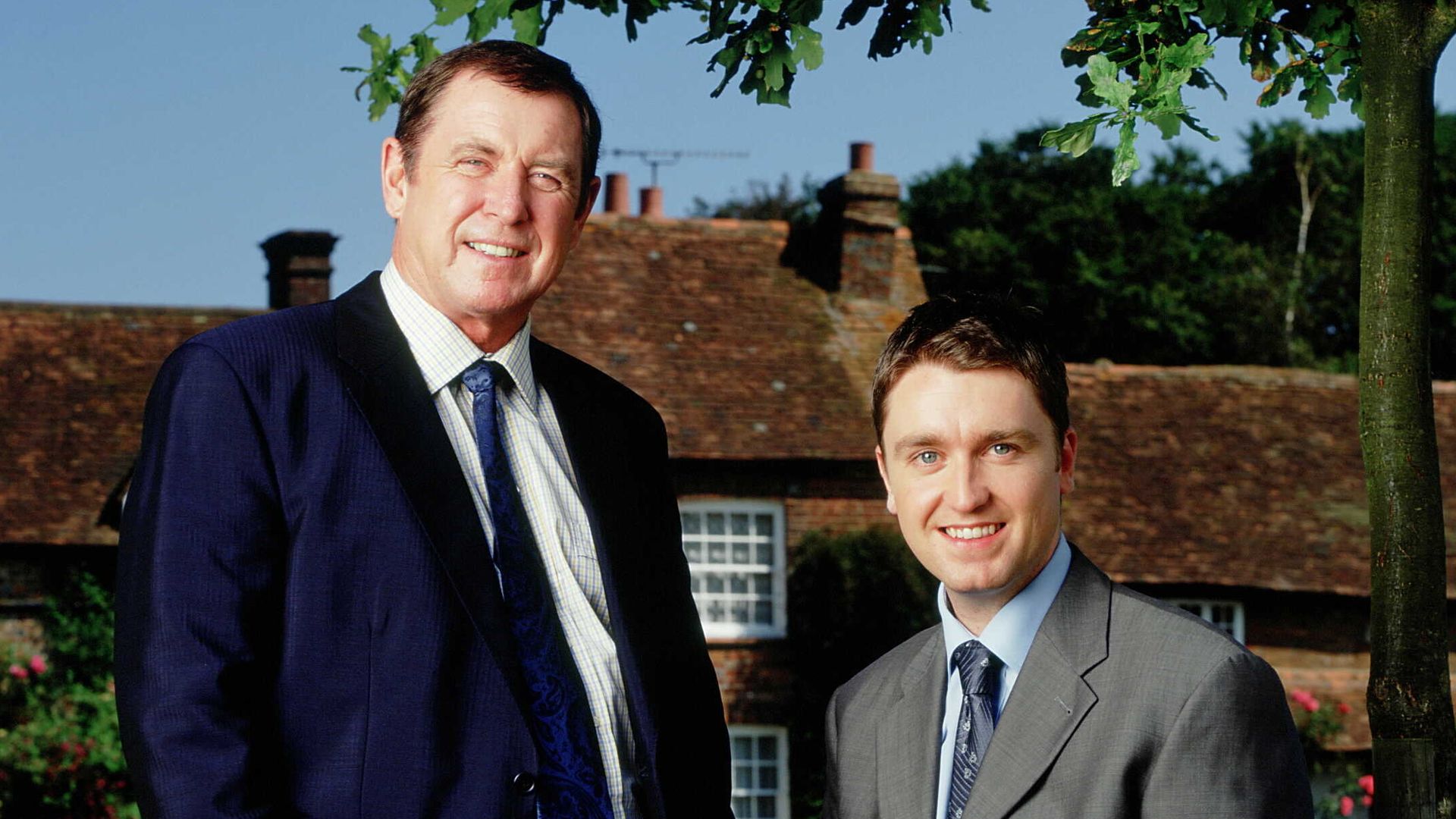 two actors in suits standing on grass outside village house