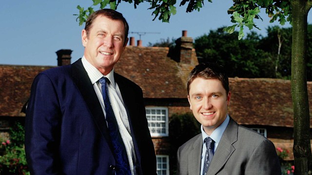 two actors in suits standing on grass outside village house