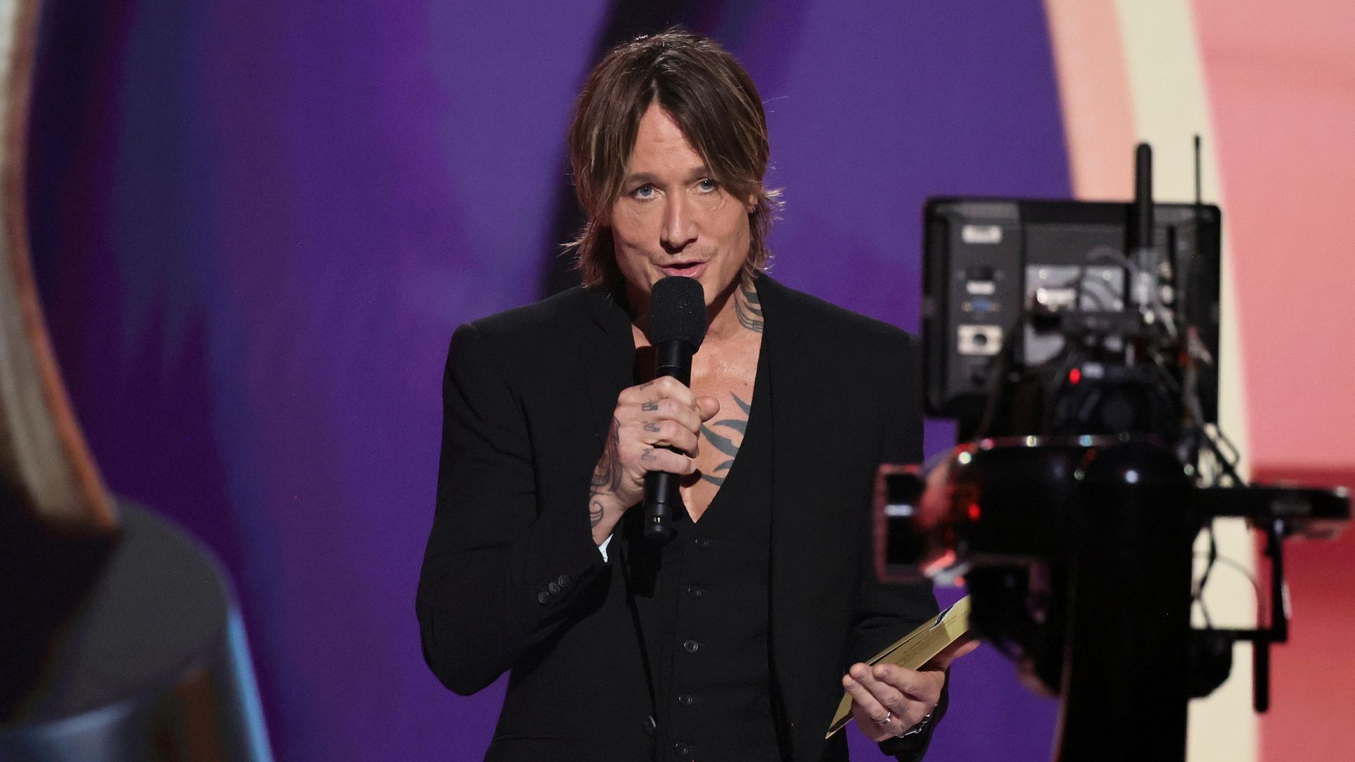 Keith on stage with a mic and awards envelope, a camera and teleprompter are in front of him