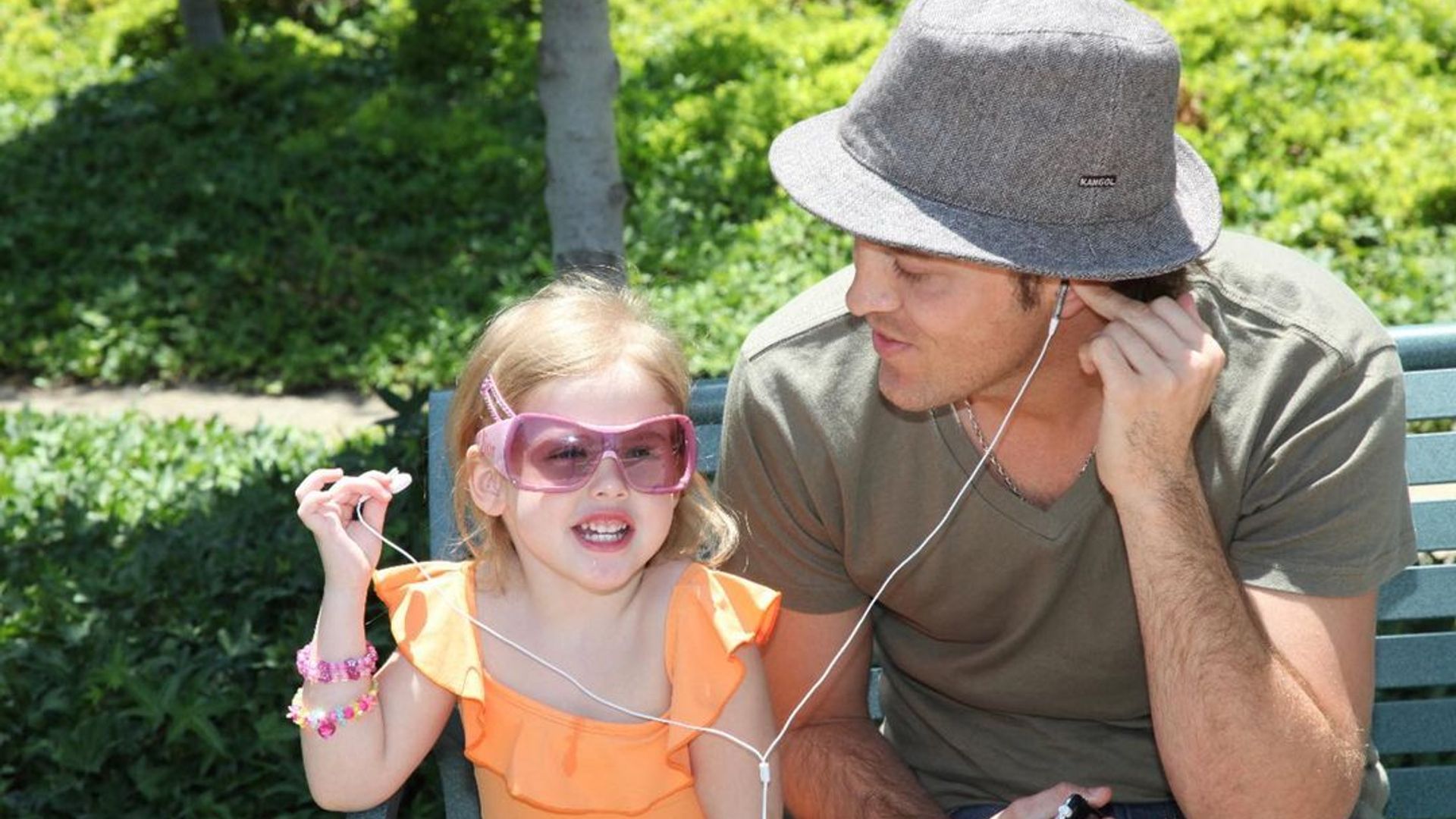 A young Dannielynn sits next to her dad on a bench in a park, sharing headphones with him