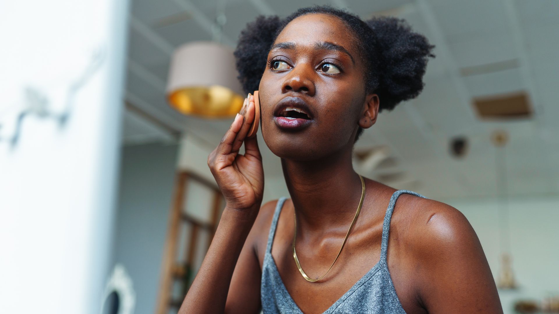 Young multiracial woman applying foundation on face with sponge while standing in front of mirror trying to hide skin problems