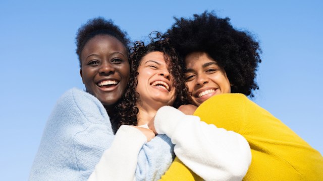 Three cheerful multi-ethnic young women embracing on blue background and two of them looking at the camera. Happy friends smiling