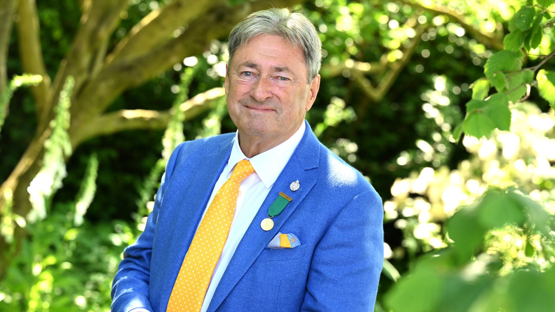 Alan Titchmarsh in a blue suit and yellow tie stands in front of trees for headshot photo for the Chelsea Flower Show