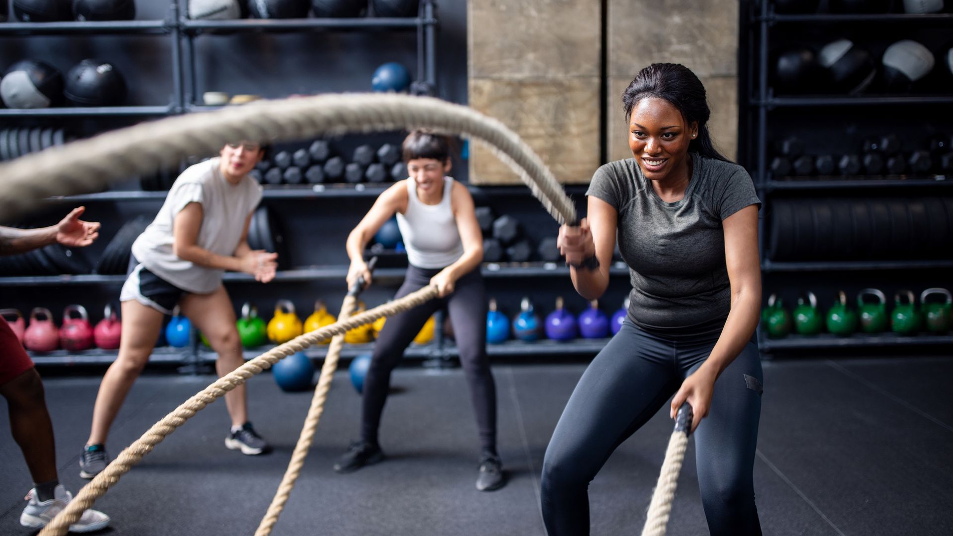 Female fitness enthusiasts engage in a vigorous battle ropes workout, supported by friends cheering them on at a cross-training gym.