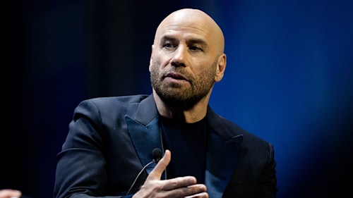 John Travolta wearing a navy suit and talking on stage in 2019