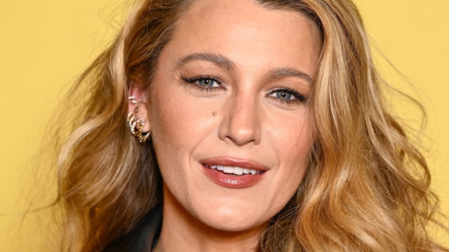 Blake Lively smiles on the red carpet in a black leather jacket dress with voluminous waves and radiant makeup, attending the premiere of A Simple Favour 2.