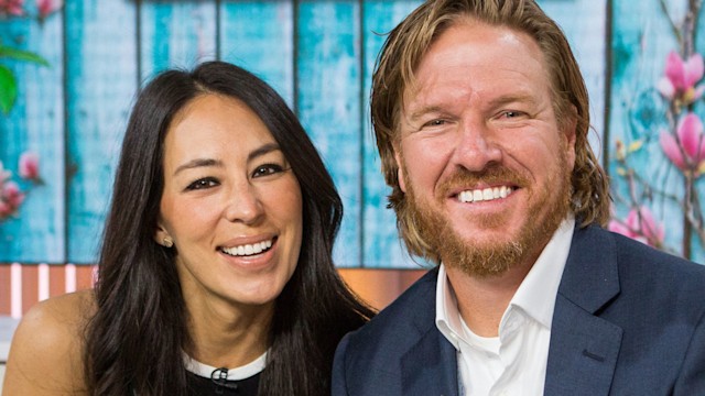 Chip Gaines in a blue suit and Joanna Gaines in a black dress smiling on TV