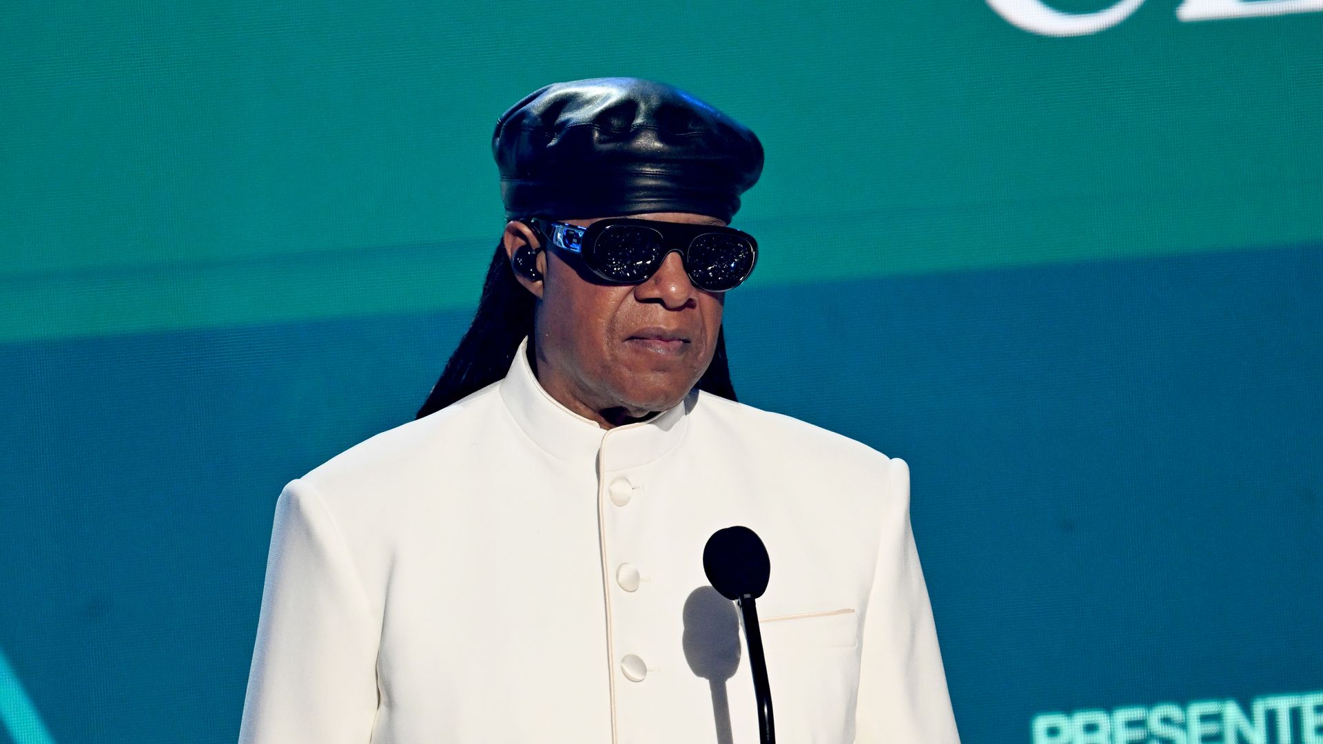 stevie wonder in white jacket and black hat on stage in front of microphone