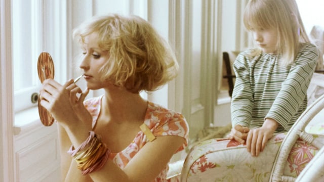 Model Chris Royer seated on the floor in front of window applying makeup with a handheld mirror, a child standing behind her and holding up a seat cushion. She is wearing Lilly Pulitzer's orange and white bird-patterned Dacron polyester and cotton pajama set of capsleeve T-shirt and pants. Accessories: bangles by Lecour-Johnson. Makeup by Sandra Linter. Hair by Harry King of Cinandre.