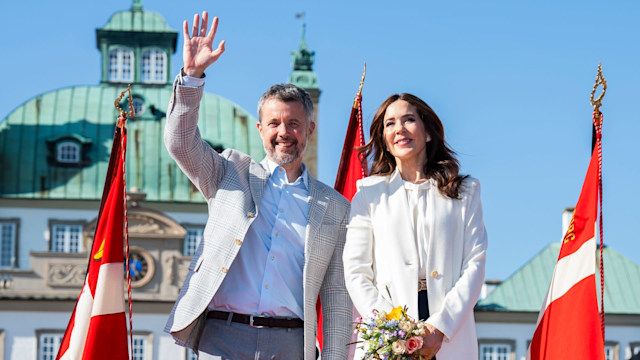 King Frederik waving and standing with wife Queen Mary who holds a bunch of flowers