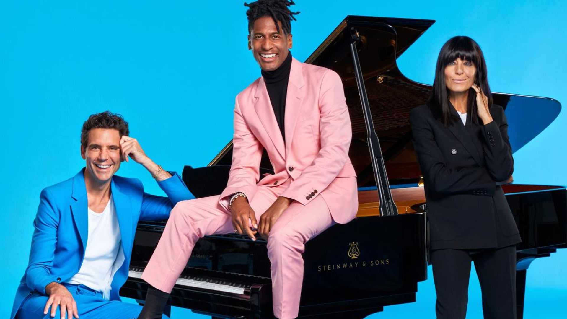 Three TV presenters posing with piano against blue backdrop