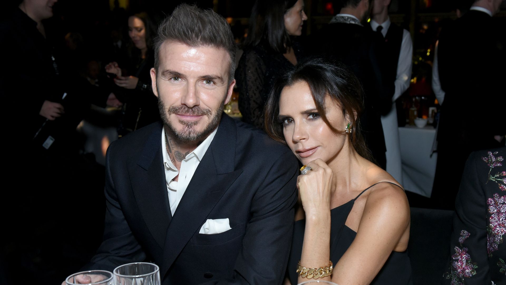 David and Victoria Beckham sat at a table at an event, smiling at the camera