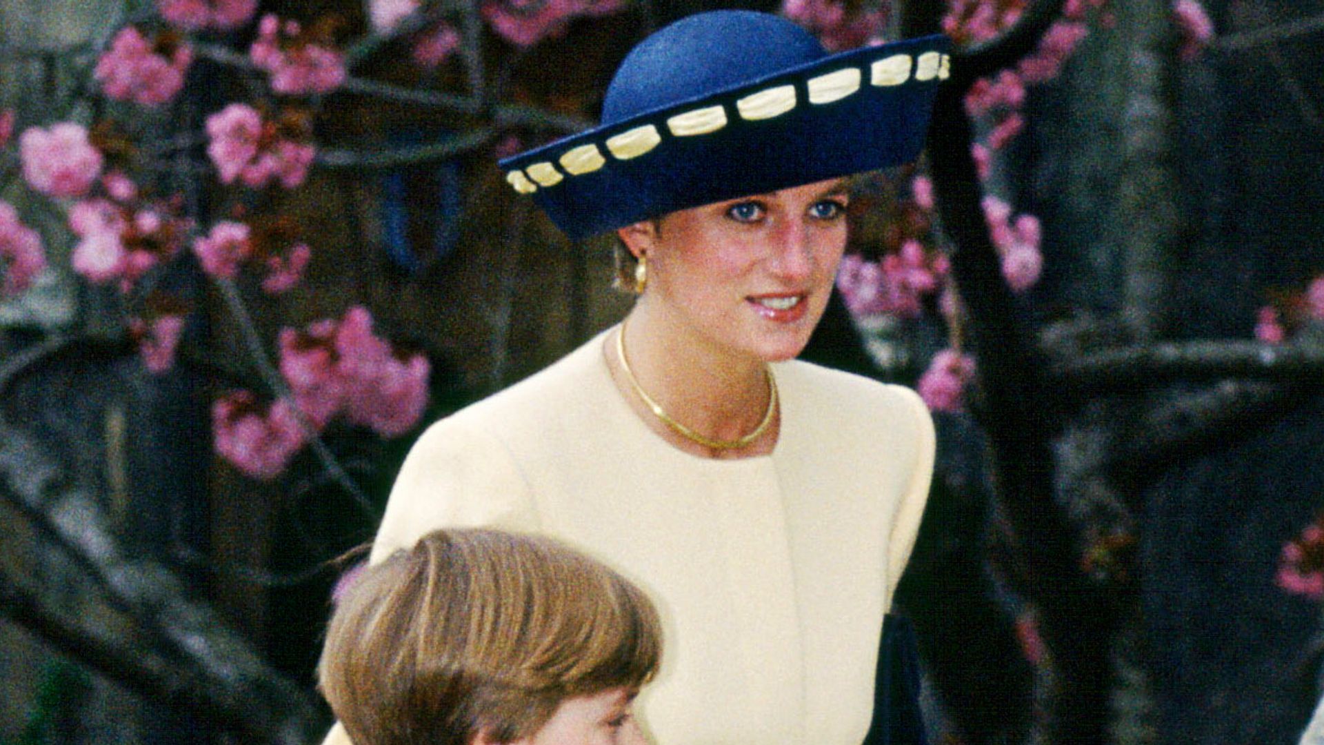 Princess Diana walks up the steps with Prince William and Lady Louise Windsor, dressed in her yellow pleated suit and navy heels, framed by pink cherry blossoms and Easter boxes in hand.