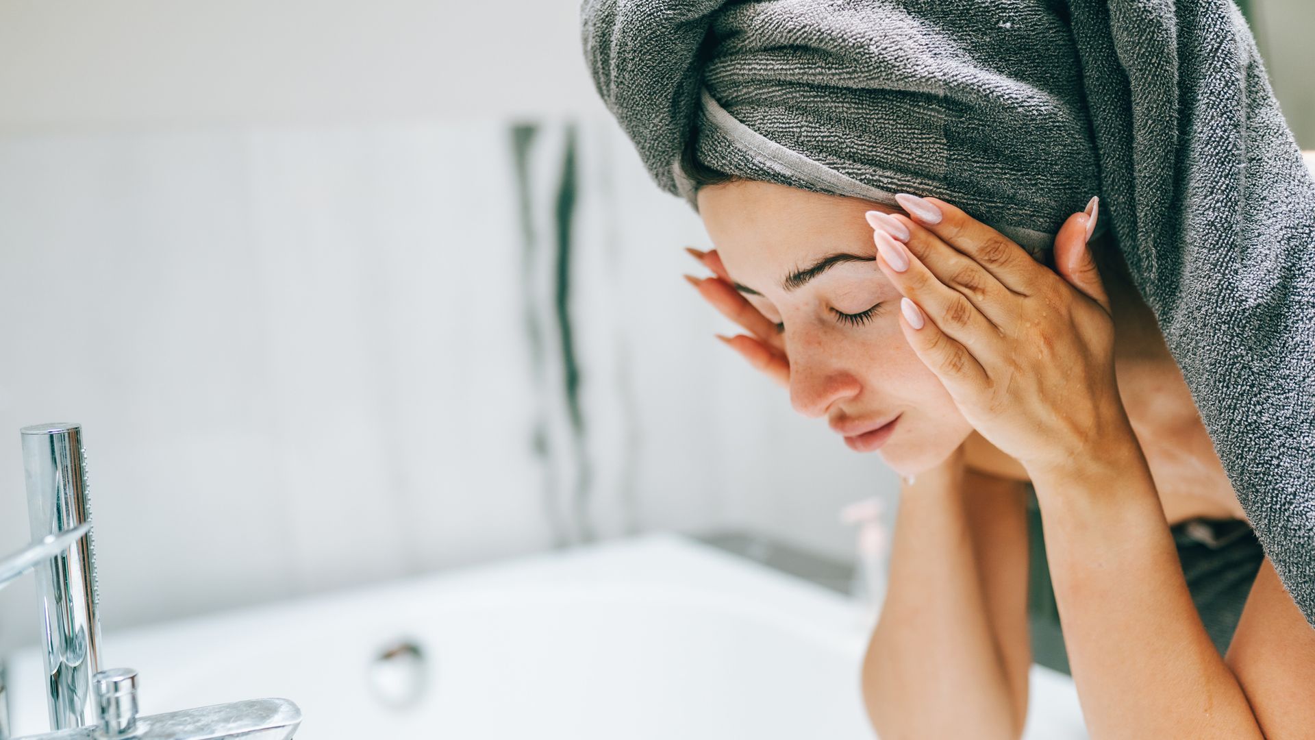 Woman caring for skin: woman in bath towel washes face early morning. She stands in front of bath, gently cleansing and applying skincare products, feeling rejuvenated