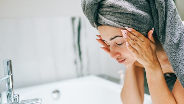Woman caring for skin: woman in bath towel washes face early morning. She stands in front of bath, gently cleansing and applying skincare products, feeling rejuvenated