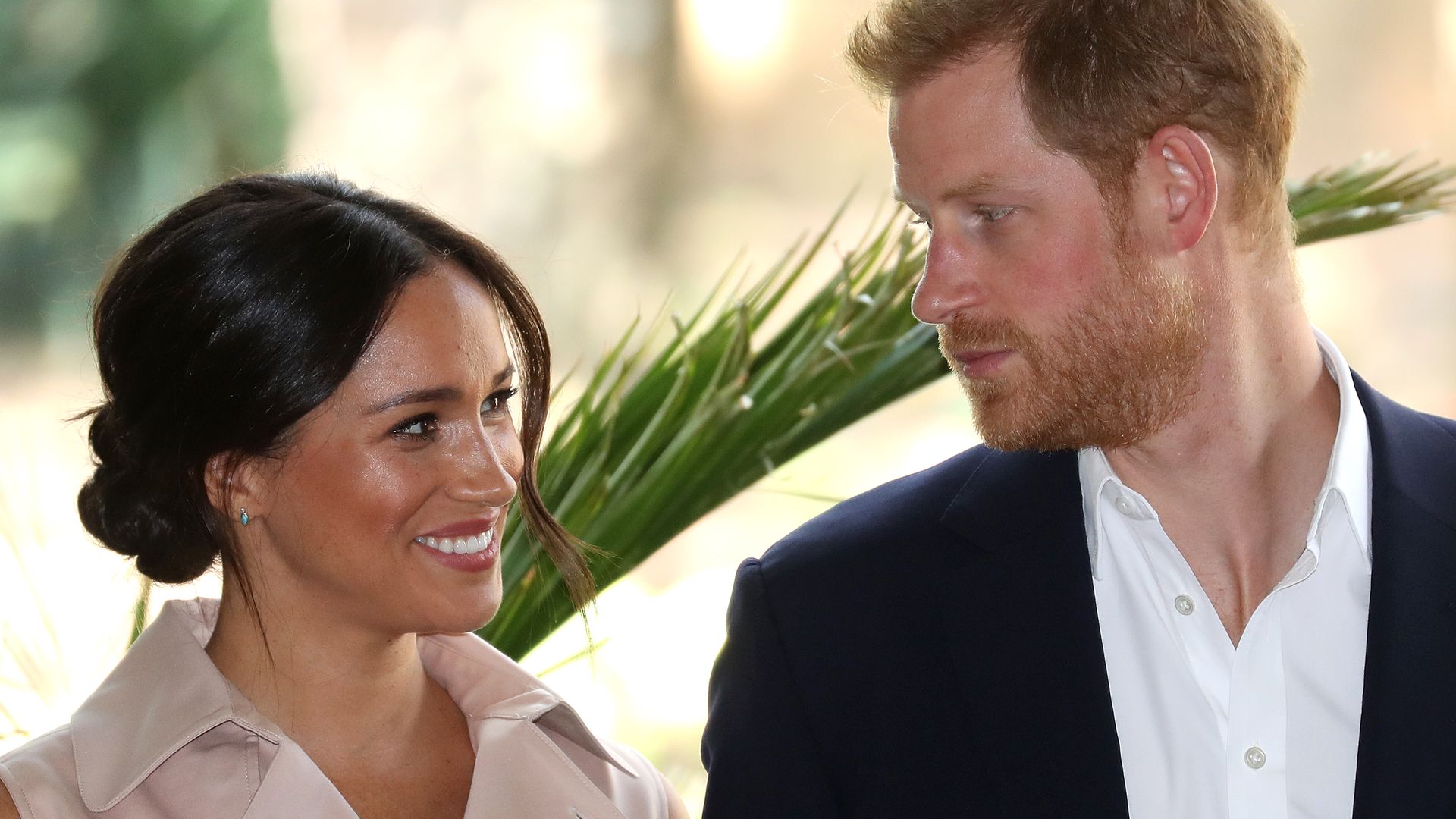 Prince Harry in a suit looking at Meghan Markle in front of a palm tree