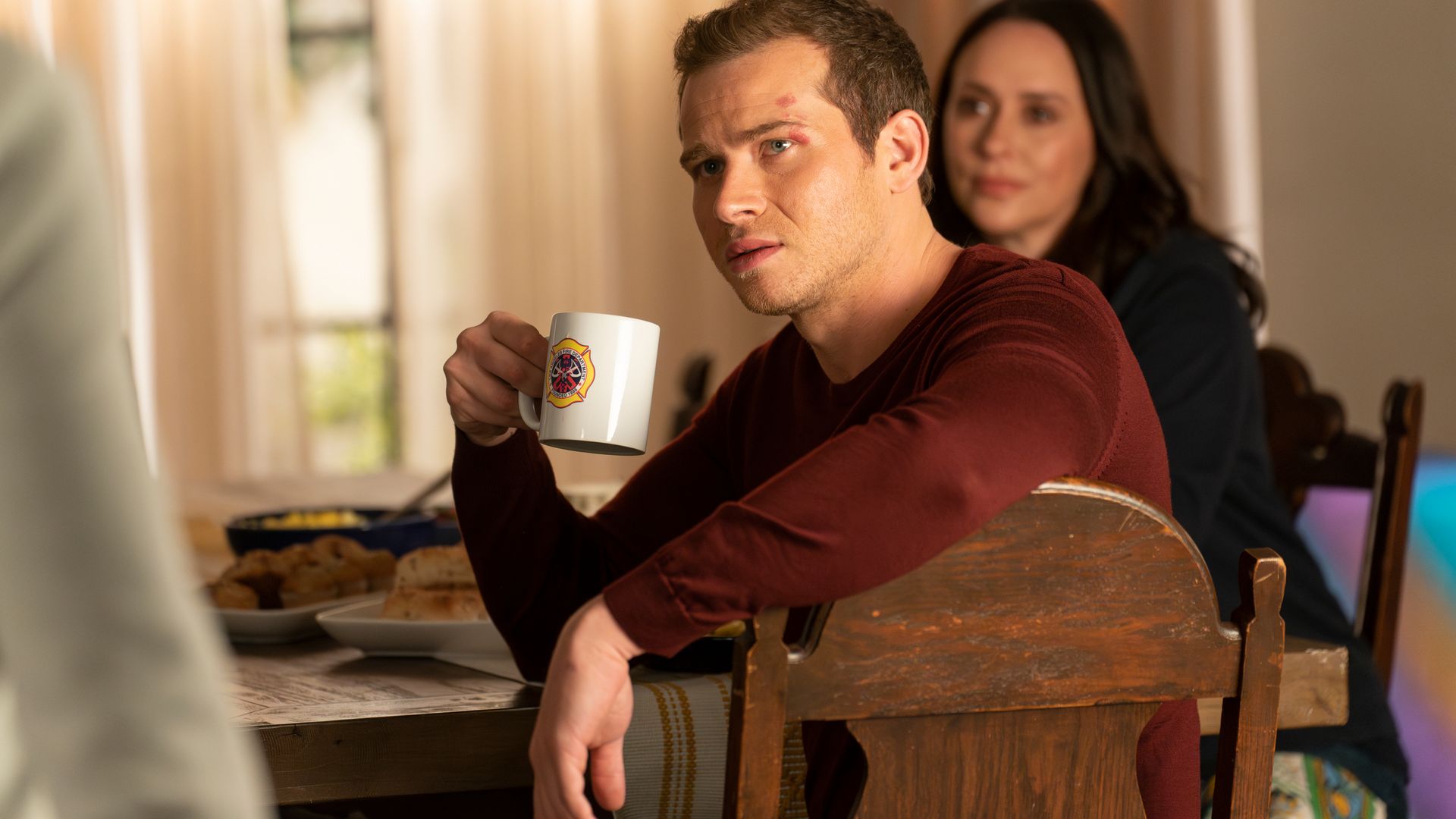 Oliver Stark holds a mug as he sits at a dining table in a still from 911

