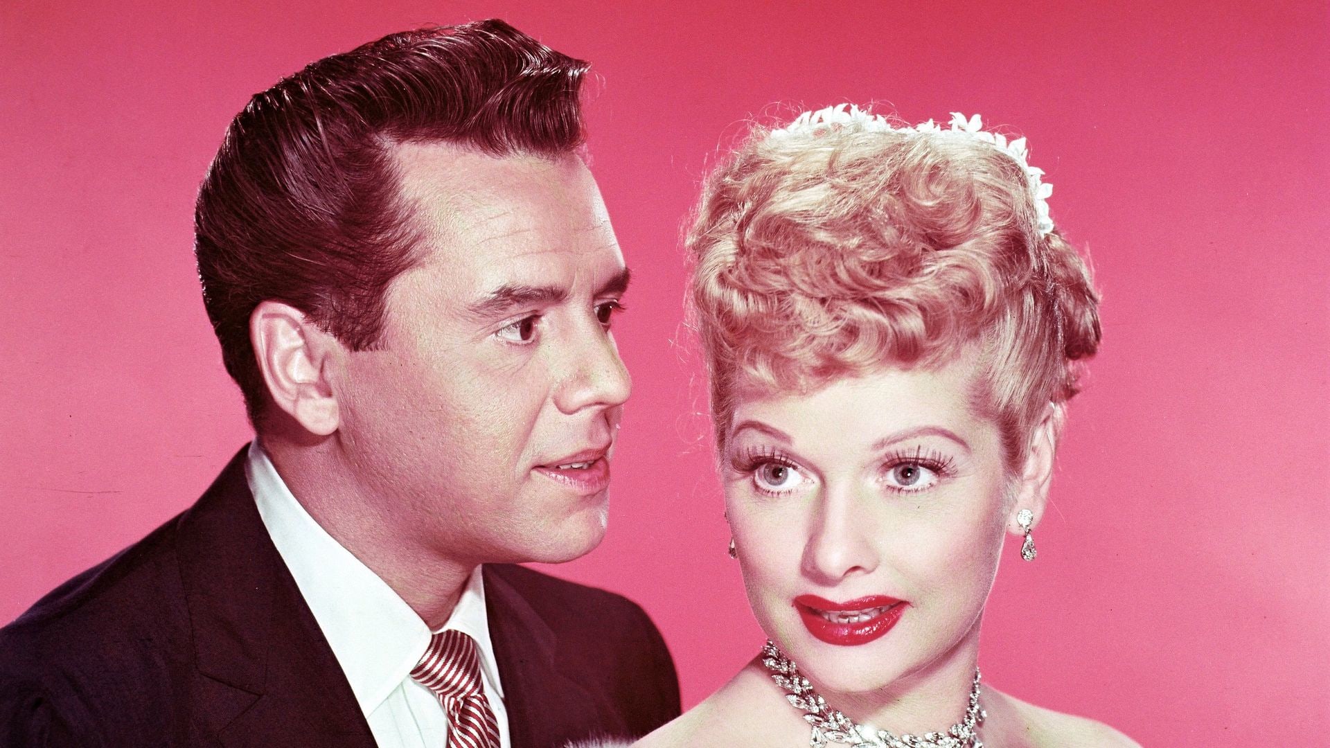 Desi Arnaz (1917-1986), Cuban musician and actor, with Lucille Ball (1911-1989), US comedian and actress, wearing a grey fur stole, in a studio portrait, against a red background, circa 1955.