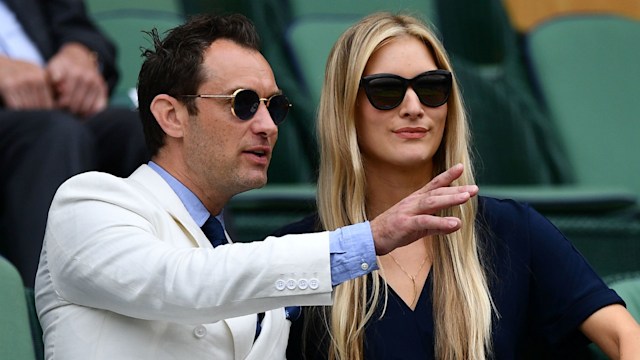 Jude Law and his wife Phillipa Coan sat at Wimbledon, both wearing sunglasses and looking to the left, Jude is pointing out to the left