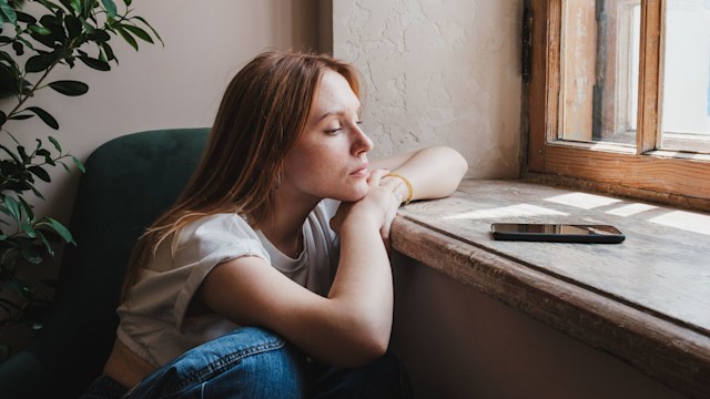 Upset redhead teen girl sitting by window looking at phone waiting call from boyfriend, feeling sad and depressed teenager looking at smartphone wait for message. Social Media depression in teens