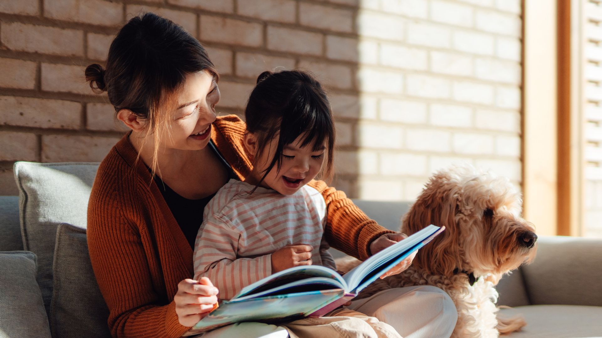 Cute Asian little girl sitting on her mother's lap, reading a book, enjoying story time, relaxing on the sofa at home with their dog. Healthy family relationship. Happy loving family.