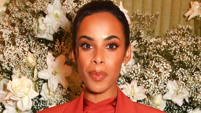 Rochelle Humes in a red feathered suits in front of a wall of white flowers