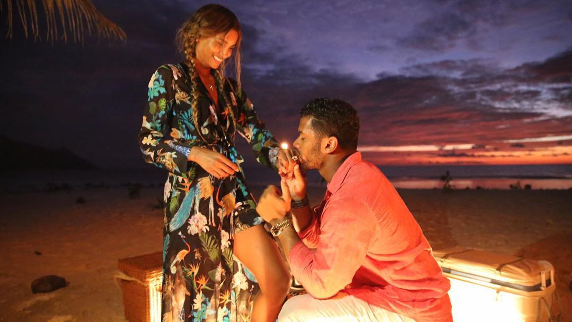 Russell kneeing on a beach, holding Ciara's hand and kissing it after he proposed, she is smiling