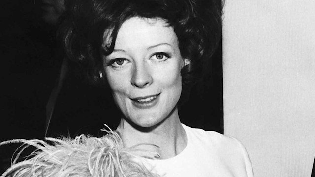 In this image, Maggie Smith is dressed in a glamorous, white gown adorned with feathers. The feathers cover her shoulders and cascade down her arms, adding dramatic flair to her look. She is holding a box, with a soft smile on her face, appearing graceful and poised. The scene captures her in an award ceremony setting, embodying Hollywood glamour with a touch of fun.